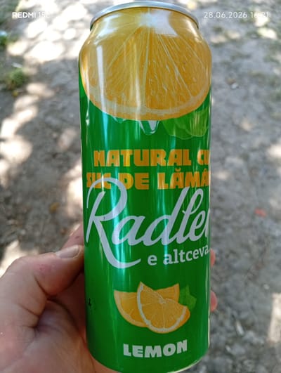 A hand holds a green aluminum can with a yellow and white label featuring a graphic of sliced lemons and text. The text on the can is in Romanian and reads "NATURAL CU SUCE DE LAMA" which translates to "NATURAL WITH LEMON JUICE" and "Radler e altceva" which translates to "Radler is something else." Below that, "LEMON" is written in English. The can is depicted outdoors in what appears to be a shaded area with a dirt or gravel ground. The background shows dappled sunlight and shadow, suggesting trees or foliage overhead. The timestamp on the image indicates the photo was taken on June 28, 2026, at 16:21.