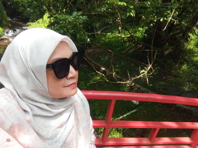 In Durin Serugun, Indonesia, a woman wearing a white hijab and dark sunglasses is captured in a selfie. She is positioned on a red bridge, with a lush green forest and a stream visible in the background. The scene is bathed in natural light, creating a vibrant and serene atmosphere.