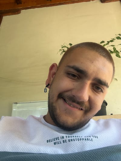 A close-up portrait of a young man, rotated 90 degrees clockwise, captures him smiling warmly at the camera. He has a short buzz cut, prominent dark eyebrows, a mustache, and a neatly trimmed beard. His white, textured t-shirt displays the motivational text, oriented vertically, which reads: "BELIEVE IN YOURSELF AND YOU WILL BE UNSTOPPABLE." Part of a grey garment is visible on his right side, possibly worn over the white shirt. A distinctive black earring with a miniature razor blade charm hangs from his left ear. The background features a plain, light yellow wall, with a wooden beam or frame visible on the far left. Green foliage, resembling a vine, is sparsely distributed in the upper left and behind his head. The natural lighting suggests a daytime setting. This image was captured in Staro Selo, Bulgaria.