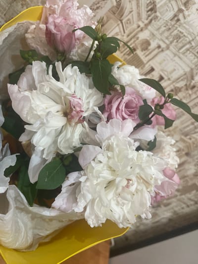 A bouquet of pink and white flowers, possibly peonies and roses, is presented in a bright yellow gift bag, with white tissue paper peeking out. The bouquet is arranged in a way that some of the petals are slightly wilted, suggesting it may not be freshly cut. The background features a textured wallpaper with a pattern that resembles classical architecture, possibly from a building in Gukovo, Russia. The lighting is natural, coming from an unseen source, and casts soft shadows on the flowers and the background. There are no people or animals visible in the image. The scene appears to be indoors, with the bouquet placed on a surface that is partially visible at the bottom of the frame, which is likely a table or shelf.