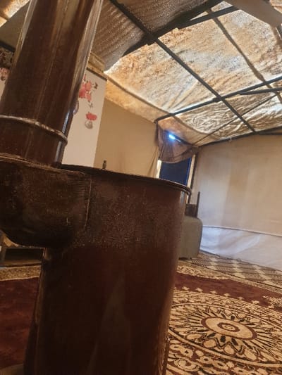 The image displays the interior of a modest living space, likely within a temporary structure in Killi, Syria. A large, dark brown pipe or part of a stove dominates the foreground, running diagonally across the frame. To the right, a section of a patterned rug with a circular, medallion-like design in shades of red and beige is visible on the floor.
The ceiling above is constructed from a translucent, light-colored fabric or plastic material stretched over a framework of dark metal bars, indicative of a tent or a makeshift shelter. In the background, a bright blue light emanates from a small opening or window, suggesting daylight filtering in. Below this light source, a white object adorned with colorful floral patterns, possibly a refrigerator or a cabinet, is partially visible. No people are present, and no specific activity is occurring. The overall impression is that of a functional, yet humble, indoor environment. There is no visible text in the image.