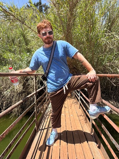 A red-haired, bearded man wearing a blue t-shirt, brown pants, and light blue sneakers stands on a wooden and metal bridge, holding a water bottle in his left hand. He also has sunglasses and a strap across his chest. Reeds are visible on the left of the frame. Water can be seen between the metal bars of the bridge, and wooden planks form the walkway. The location is Misgav Regional Council, Israel.