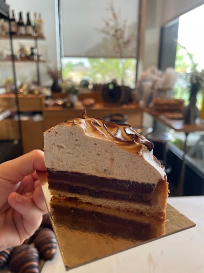 A close-up view depicts a slice of multi-layered dessert held by a light-skinned human hand, positioned in the foreground of an upscale commercial establishment in Nes Ziona, Israel. The dessert slice features a prominent, torched meringue topping with browned peaks, layered above two distinct dark, glossy chocolate or caramel fillings separated by a thinner dark brown layer, all resting on a light brown pastry crust. The slice is supported by a reflective, gold-colored rectangular cardboard base. The background, largely out of focus, reveals an interior space with wooden shelves displaying various bottled products and jars, along with counters containing blurred items. Large windows on the right side of the frame offer a view of indistinct outdoor greenery and structures, indicating natural light illuminating the scene.