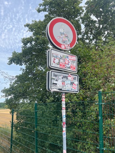 The image depicts a street sign in Rheinbach, Germany, adorned with numerous stickers. The signpost is surrounded by lush green trees and foliage, with a chain-link fence in the foreground and a field of dry grass to the left. The sky is partly cloudy. The sign itself appears to be a no-parking sign, but it is heavily obscured by stickers. Some of the visible text on the stickers includes "ULTRAS", "ACAB", "UNSER LEBEN", and "HANGT AN DIR". The overall impression is one of a rural or suburban roadside setting.