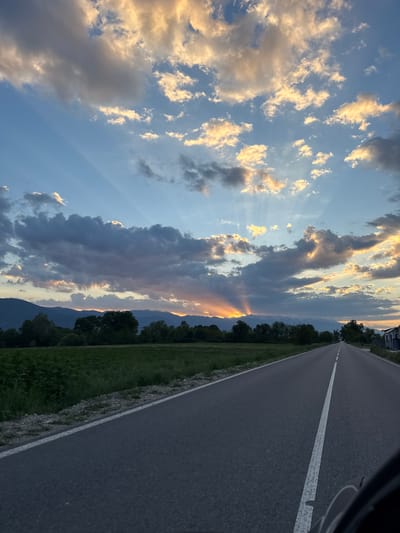 The image shows a rural road near Hvostiane, Bulgaria, during sunset. The asphalt road has a single white center line and a white shoulder line on the right side. Adjacent to the road is a narrow strip of grass and trees, followed by mountainous terrain in the distance. The sky occupies a significant portion of the image, filled with clouds illuminated by the setting sun. The sunlight is visible in rays passing through gaps in the clouds. No people are visible in the image.