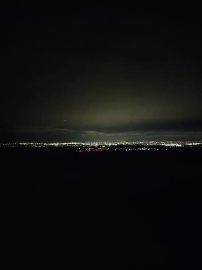 The image displays a panoramic night view of the city of Balsha, Bulgaria, captured from an elevated perspective. The lower half of the frame consists of an undifferentiated dark expanse, devoid of discernible features. Above this, a horizontal band of numerous, scattered artificial lights, primarily yellow-orange and white, delineates the urban landscape. These lights appear as blurred points, indicating distance. The upper portion of the frame is occupied by a dark, overcast night sky with visible cloud formations, including a prominent darker cloud band just above the city lights and lighter, hazier clouds higher up. A single, distinct, bright white point of light, consistent with a star or planet, is visible in the upper-mid section of the sky. No individuals, specific actions, or interactions are observable within the scene.
