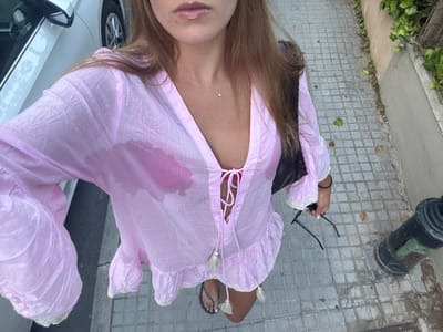 The image displays a female individual from a high-angle perspective, standing on a tiled sidewalk in Cas Català-ses Illetes, Spain. The individual is wearing a wet, light pink, sheer long-sleeved cover-up with a deep V-neck and lace-up front, through which a bright pink bikini top is visible due to moisture saturation on the fabric. She is wearing dark flip-flops. A black shoulder bag is slung over her left shoulder, and she holds black sunglasses in her right hand. A delicate silver necklace with a small pendant is visible on her neck. To the left, a section of a white parked vehicle is visible. To the right, a wall covered in green ivy or similar foliage is present, along with a black cylindrical street bollard or post in the foreground. The sidewalk is paved with grey square-patterned tiles, and a few dry leaves are scattered on its surface.
