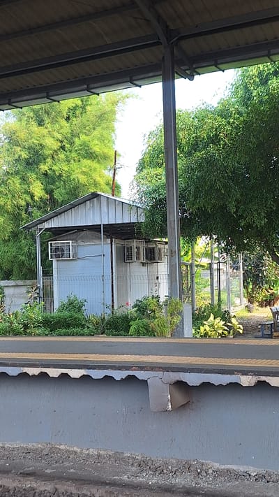 This image depicts a small, light-colored building with a metal roof, situated amidst lush greenery and trees, suggesting a semi-rural or park-like setting within the city of Karawang, Indonesia. The building appears to be a utility structure, possibly a guardhouse or storage unit, with multiple windows equipped with air conditioning units. The foreground shows a concrete platform, likely part of a public space such as a train station or park entrance, with rough gravel below. The overall scene is characterized by natural elements like dense foliage and a clear sky, juxtaposed with man-made structures. There are no people or animals visible in the image.