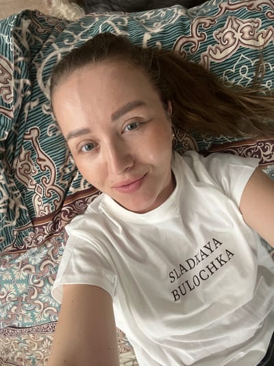 A fair-skinned woman with brown hair and blue eyes is lying on a patterned bedspread. She is wearing a white t-shirt with Cyrillic text printed vertically on the right side. The text reads "SLADKAYA BULOCHKA". The bedspread is a mix of teal, brown, and beige patterns. The location cannot be determined from the image.