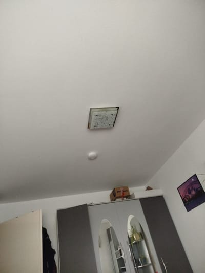 The image depicts an indoor scene, likely a bedroom or living area, in Ehingen (Donau), Germany. The perspective is looking upwards towards the ceiling.
Dominating the upper portion of the image is a plain white ceiling. Centered on the ceiling is a square light fixture with a decorative glass cover featuring an intricate pattern. Below this light fixture is a small, white, circular object, which appears to be a smoke detector.
Towards the right side of the frame, the ceiling slopes down, meeting a white wall. On this wall, a framed picture is hung at an angle. The picture shows a silhouette of the Sydney Opera House and Harbour Bridge against a purple and pink sunset or sunrise.
Below the ceiling and to the left, a large wardrobe or cabinet with grey doors is visible. The doors appear to have reflective surfaces, showing a distorted reflection of plants and shelving within. To the left of the wardrobe, a plain, light-colored cabinet door is partially open, revealing a glimpse of dark fabric hanging inside, possibly clothing.
On top of the wardrobe, a cardboard box is placed. The box has some red strapping and a logo with text that includes "Sport".
The lighting in the room is dim, suggesting it might be evening or the blinds are drawn. The overall impression is of a typical domestic interior. No people are visible in the image.