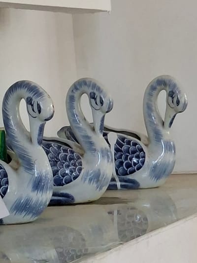 Three blue and white ceramic crane figurines are stacked vertically. The figurines are detailed with scales and have elongated necks that curve to form a hook-like head. They appear to be indoors. There is no indication of the city of Uleereuleung, Indonesia.