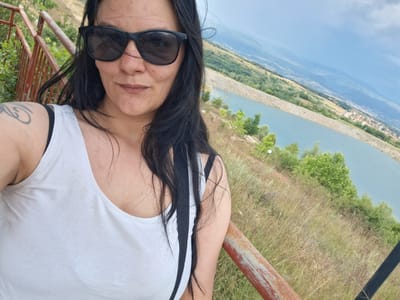 A woman with long, dark hair and sunglasses poses for a selfie outdoors. She is wearing a white tank top. Behind her, a body of water, likely a reservoir, is visible, with hills and a town in the distance. The location is identified as near Dobrotino, Bulgaria. The scene appears to be a scenic overlook.