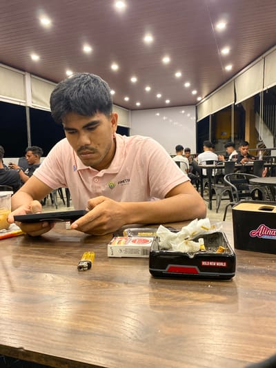 The image captures a casual evening scene at a cafe or restaurant in Keudeublang, Indonesia. The primary subject is a young man with dark hair and a light beard, wearing a pink polo shirt with "AMITR" and "syariah" visible on it. He is engrossed in his black smartphone, held in both hands, while seated at a wooden table.
On the table, an ashtray is prominently displayed, filled with cigarette butts and crumpled tissues, bearing the text "BOLD NEW WORLD." Beside it are a yellow cigarette lighter and a white cigarette pack showing the "BAT" logo. A black container with "Alinaa" written on it is also visible on the table.
In the background, several other young men are present, sitting at dark tables and chairs, engaging in conversation or waiting, suggesting a social gathering. The setting features a ceiling with multiple bright, recessed lights, indicating that the picture was taken at night or in the evening. The overall ambiance is relaxed and social.