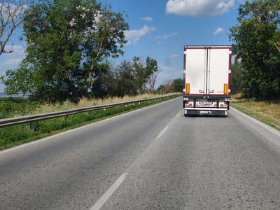 The image shows the rear view of a white semi-trailer truck driving on a paved road. The truck has red trim around the doors and yellow reflective markers. The license plate reads "RV 560VK" and below that is the brand name "SCHMITZ" and "The trailer company." The truck is in the right lane, and the road curves gently to the left.
The setting is a rural area with green trees and foliage on both sides of the road. A metal guardrail runs along the left side of the road, separating it from a grassy embankment. The sky is blue with scattered white clouds, indicating it is daytime with fair weather. The sun appears to be high in the sky, suggesting it is likely midday. The road surface is asphalt and appears to be in good condition, with white lane markings.
The perspective is from a vehicle following the truck, giving a sense of movement along the road. No people are visible in the image. The overall scene depicts a typical highway journey through a countryside landscape. The location context provided is Totleben, Bulgaria, which aligns with the rural and European-style road features.