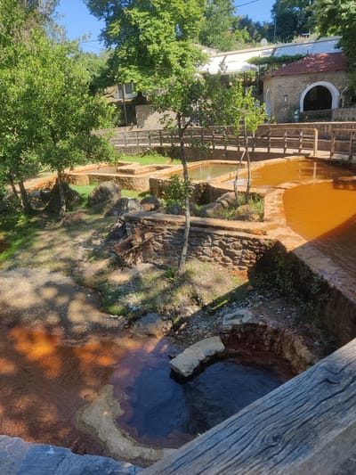 This is an outdoor, eye-level, full shot of a scenic natural setting that appears to be within the municipality of Evrymenon, Greece. The foreground is dominated by a rustic, weathered wooden railing that partially obstructs the view of several tiered pools of water. These pools vary in color and depth; some contain a reddish-brown, mineral-rich water, while others hold darker, possibly deeper water. The pools are bordered by rough-hewn stone structures and natural rock formations. Lush green trees with dense foliage provide shade and frame the upper part of the image, with their dappled shadows cast across the ground and water. In the background, a traditional stone building with a red-tiled roof and an arched entryway is visible, suggesting a possibly established or utilized site. A wooden walkway or bridge is present, connecting different sections of the area. The overall environment is one of natural beauty, with a clear blue sky overhead and abundant greenery. There are no people or animals visible in the image.