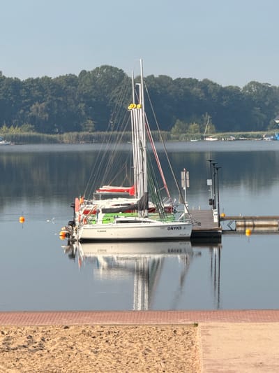 The image depicts a serene waterscape with sailboats docked at a marina on a clear, sunny day. In the foreground, a sandy beach borders a paved walkway with brick accents, suggesting a recreational area. The middle ground is dominated by the lake and the docked sailboats. One prominent sailboat, labeled "ONYKS," is tethered to a wooden dock. The sailboat has multiple masts and rigging, and a red cover is visible on its boom. Other sailboats are also docked, with some displaying green and red covers. Several orange buoys float on the calm water, marking the area. In the background, a dense line of trees forms the shoreline, with a clear blue sky above. The water reflects the sky and the sailboats, creating a symmetrical and peaceful atmosphere. The location is identified as Sława, Poland. There are no people visible in the image.