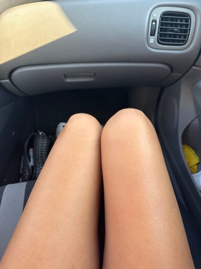 This is a close-up, medium shot, eye-level, indoor photograph of a person's bare thighs, taken from a first-person perspective. The person is seated in what appears to be the passenger seat of a car, with the dashboard and part of the car interior visible. The lighting suggests a daytime setting, with sunlight casting a shadow across the top left of the dashboard. The car's air vent and a glove compartment are visible in the background. The person's skin is tanned. A black patterned bag is on the floor of the car to the left. The image does not provide enough information to determine the specific location within Sagaidac, Moldova.