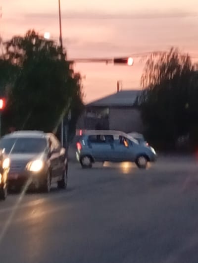 The image shows a street scene in Salar, Uzbekistan, at dusk. A light-colored car is in motion on the right side of the frame. Another car is in the foreground on the left side. The taillights of the car on the right are illuminated. The traffic lights are visible. Buildings are in the background and trees border the street.