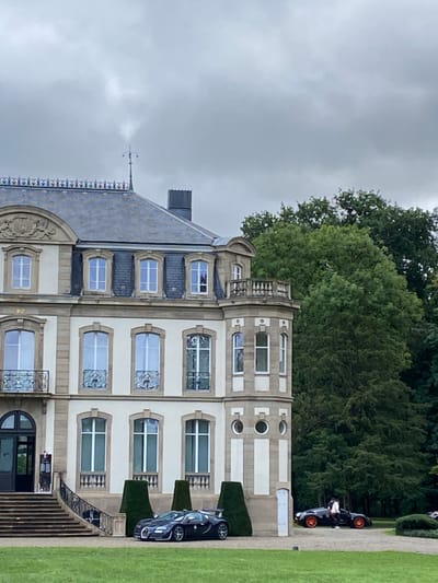 An image captured in Dorlisheim, France, displays a large, ornate building with a well-maintained lawn in the foreground and dense trees in the background under an overcast sky.

The building, reminiscent of a château, features a light-colored facade with tan stone accents and a dark grey slate roof. The roof is adorned with a decorative railing and a central spire. Multiple dormer windows are present on the roof, alongside a visible chimney. The building's architecture includes several rectangular windows with white frames; those on the second floor above the main entrance feature decorative wrought-iron balconies. The main entrance on the ground level consists of dark double doors, accessed by a wide stone staircase with railings. The right side of the building features a multi-story octagonal or rounded tower section, complete with a small balcony on its upper level and a combination of rectangular and circular porthole windows on its lower floors. Two dark green conical topiaries are positioned near the building's facade on the lawn.

On the green lawn in front of the building, two high-performance automobiles are parked. The vehicle on the left is a dark blue or black Bugatti Veyron, positioned facing left. The vehicle on the right is a black and orange Bugatti Chiron, parked facing right. A single individual is visible near the driver's side door of the black and orange Bugatti. This person is dressed in a white shirt and dark trousers, appearing to be either entering, exiting, or standing beside the open door of the vehicle. The background on the right is dominated by a dense stand of green deciduous trees, and the sky is uniformly overcast grey.
