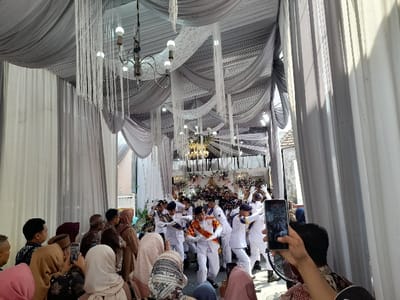 The image captures a lively celebratory event taking place in Kecamatan Gempol, Indonesia, under what appears to be a large tent or canopy. The scene is brightly lit by natural sunlight, suggesting it is daytime.
In the foreground, a crowd of people, primarily seen from behind, are actively engaged with the event. Many individuals, including several women wearing headscarves (hijabs), are holding up smartphones to record the performance unfolding before them. Their attention is directed towards the center of the frame.
The main subjects are a group of young men in the middle ground, dressed in crisp white uniforms with distinctive sashes (some orange, some blue/yellow), and dark headwear. They are performing what appears to be a synchronized dance or routine, with various poses and raised arms, indicating energetic movement.
The setting features an elaborately decorated ceiling with draped gray and white fabric, creating a festive atmosphere. Multiple chandeliers with exposed bulbs hang from above, complemented by long white decorative tassels or fringe. In the background, beyond the performers, a decorated area or stage with floral arrangements and additional lighting is visible. A glimpse of exterior buildings can be seen through an opening on the right side, confirming a semi-outdoor or tented venue. A large floor fan is also visible on the right, near the audience.
No discernible text is visible in the image. The overall impression is one of a festive community gathering or personal celebration, possibly a wedding or graduation, marked by lively entertainment and an engaged audience.