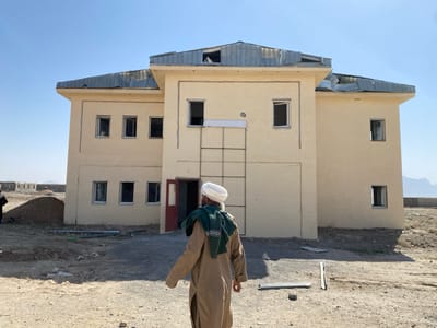 The image depicts an outdoor scene in Bar Mel, Afghanistan, on a clear, sunny day. The primary subject is a two-story, light tan-colored building with a partially damaged or incomplete silver corrugated metal roof exhibiting buckling. The building features multiple rectangular window openings on both floors, most appearing empty without glass panes. A red door, partially ajar, is visible on the ground floor of the central facade. A vertical ladder-like outline or structure is etched into the facade above the door.
In the foreground, an individual is seen from behind, walking away from the viewer towards the building. This person wears a tan-colored traditional garment, a white turban, and a green patterned scarf draped over their shoulders. The ground in the immediate foreground is unpaved, dry, and dusty, with scattered debris including two rectangular dark metal sheets or panels. To the left of the building, a large mound of loose earth or sand is visible. In the far left background, several smaller, incomplete structures or buildings are discernible. The distant background features a flat, arid landscape with faint outlines of mountains under a clear blue sky. No specific interactions are observed.