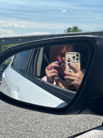 A person is taking a selfie in the side-view mirror of a car. The person has long, light brown hair and bright blue fingernails. They are wearing a pink shirt. They are holding a smartphone with a light-colored case that has cartoon characters on it. A pink ring is visible on one of their fingers. The car is white and appears to be in motion, as suggested by the blurred background of asphalt and a guardrail. The sky is blue with scattered clouds. The location in the city of Крутобереговый, Russia is not evident from the image.