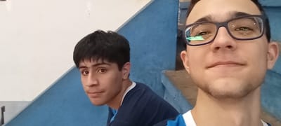 The image shows two young men. The man on the right is wearing blue framed glasses, and a white shirt with a blue trim. He has a light beard and is smiling. The man on the left is looking at the camera, and is wearing a blue shirt with a white collar. He is wearing a dark blue shirt with a white collar. Both men are facing the camera. The background is a blue wall with a white wall in the distance. The image was taken in Encarnación, Paraguay, at latitude -27.332 and longitude -55.871.  The location is likely inside a gymnasium or other building.