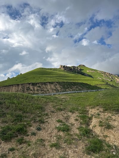 The image depicts a scenic view of a steep, grassy mountainside with a road cut into its lower slope. The mountain is covered in vibrant green grass, with rocky outcrops visible near the summit. A guardrail runs along the edge of the road, suggesting it is a route for vehicles. The foreground shows a drier, more arid terrain with sparse vegetation and loose soil. The sky above is dramatic, filled with a mix of white and grey clouds against a blue sky. The lighting suggests it is daytime, possibly late afternoon given the shadows. The scene is likely located in the natural landscape surrounding the village of Verkhnyaya Mara, Russia, known for its mountainous terrain. There are no people visible in the image.
