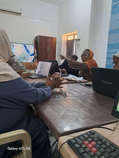 The image shows a meeting room in Karima, Sudan (latitude: 18.54, longitude: 31.831). Four people are seated around a table. The person seated at the far end of the table is wearing a blue jacket. The person in the foreground is wearing a grey jacket and is seated on a chair.  There are multiple laptops on the table. One of the laptops is open and has a map on its screen. There is a computer keyboard on the table next to a laptop. The meeting room has white walls and a wooden door. The windows are covered with a white curtain. There is a brown wooden cabinet on the left side of the room.  A blue poster with white text is on the wall. The wall is also decorated with a red flag.  The people at the meeting appear to be looking at a laptop.