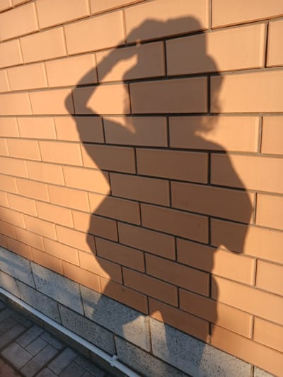 A full-body shadow of a person is cast on a light orange brick wall. The person appears to be standing and touching their head with their right hand. The shadow's left arm is bent at the elbow and held away from the body. The shadow's torso is visible, and it seems the person is wearing clothing. The bottom of the wall is adorned with a darker gray stone base, and a white pipe runs horizontally along the base. Below the wall, a paved walkway of gray rectangular stones is visible. The lighting suggests it is either early morning or late afternoon, as the sun casts a long shadow. The scene is located outdoors in Maslova Pristan', Russia.