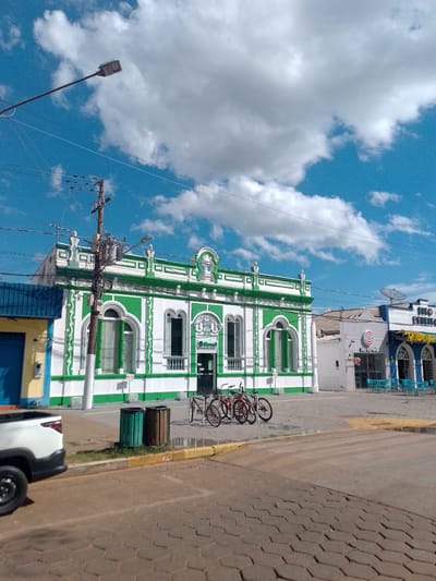 In Cáceres, Brazil, a street scene unfolds under a bright, blue sky dotted with fluffy white clouds. A white pickup truck is parked on the left side of the image, its front visible. In front of a vibrant, green and white building with ornate architectural details, three bicycles are parked. The building appears to be a commercial establishment, with signage visible. Adjacent to it, another building with blue and yellow accents is partially visible. The ground is paved with hexagonal cobblestones. Two trash receptacles, one green and one brown, stand near the bicycles. A lamppost extends from the upper left corner of the frame, and electrical wires are strung across the sky. In the background, further buildings and outdoor seating areas suggest a town square or commercial district.