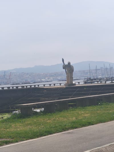 This image captures a serene daytime view of the coast in Moaña, Spain, under a hazy sky. In the foreground, a paved path borders a vibrant green grassy slope. Beyond this, a series of aged, tiered concrete structures, possibly remnants of a terraced seating area or old dock, extend towards the water.

Dominating the midground is a light-colored stone sculpture of a robed figure, holding a long staff or spear, gazing towards the right side of the frame. This figure stands on a concrete platform or pier that parallels the coastline. The calm waters of what appears to be a bay or ría are dotted with numerous moored sailboats, their masts reaching towards the sky. Further out in the water, distinct rectangular platforms, characteristic of oyster or mussel rafts (bateas), are visible.

The background reveals a bustling coastal city built into the slopes of distant hills or mountains. Densely packed buildings climb upwards, with the hazy atmosphere lending a soft, almost diffused appearance to the urban landscape. On the far left, a prominent red industrial crane can be seen within the city, suggesting port activities. No discernable text is visible in the image. The scene conveys a peaceful, observational quality of a maritime landscape.