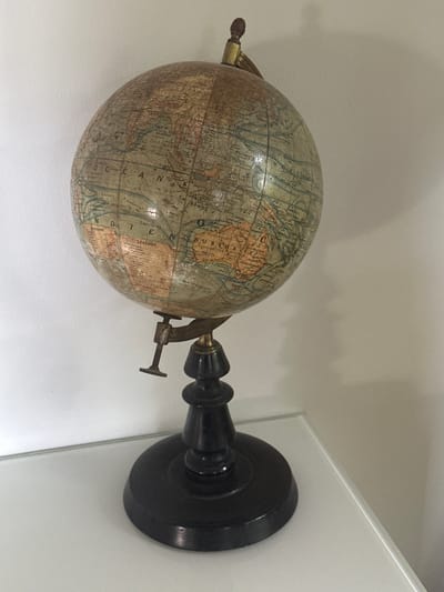 A vintage globe rests on a dark wooden base, which sits on a white surface. The globe displays maps of the world with muted colors, indicating its age. The stand features an adjustable brass clamp that holds the globe. The background is a plain, light-colored wall. The scene is indoors, with the lighting suggesting it's daytime. There are no people or animals visible in the image. The globe is the central subject, showcasing faded geographical details and labels.