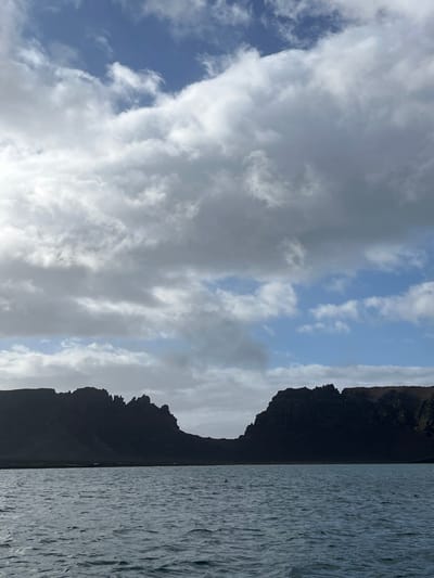 A scenic landscape features a body of water with rippled surface on the right, possibly a lake or sea. A dark, rugged mountain or cliff dominates the center, its silhouette sharp against the sky. The sky on the left is filled with billowing white clouds against patches of blue, creating a sense of depth and atmosphere. The image is oriented vertically, capturing the natural beauty of a mountainous waterscape.