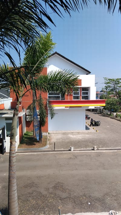 An exterior, eye-level, medium shot captures a modern, two-story building with a red and white facade, accented by a red and yellow striped overhang. The building appears to be a commercial establishment, possibly a convenience store, situated in Bojongloa, Indonesia. A palm tree with a thick, segmented trunk stands prominently in the foreground, its fronds partially obscuring the upper portion of the building. To the right of the building, a paved area is visible, featuring two motorcycles parked near yellow bollards. One motorcycle is occupied by a person wearing a helmet. The ground in front of the building is a gray asphalt road. The sky above is a clear, bright blue. The environment suggests a sunny day.