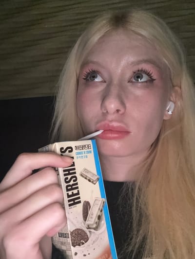An individual with blonde hair and fair skin is depicted in a close-up, illuminated by an unseen light source, located in the city of Namyangju-si, South Korea. The person's eyes, light in color, are directed upwards and slightly to the right, framed by prominent black winged eyeliner and pinkish eyeshadow. Full lips are slightly parted, holding a white straw. A white wireless earbud is visible in the individual's right ear. The right hand holds a rectangular Hersheys Cookies 'N' Creme milk carton. The carton is predominantly white with a blue side panel, featuring the "HERSHEY'S" logo, text "COOKIES 'N' CREME" (쿠키앤크림), and illustrations of Oreo-style cookies and white chocolate bars with cookie pieces in milk. The background is uniformly dark and indistinct, with faint horizontal textures visible in the upper left quadrant. The individual appears to be drinking from the carton.