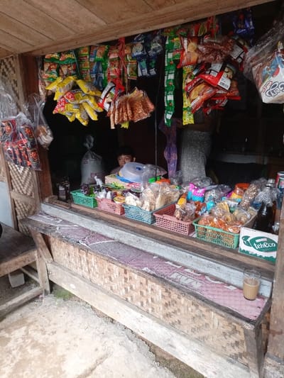 The image shows a small store in Cisewu, Indonesia, located at coordinates -7.411 latitude and 107.542 longitude. The store has a wooden frame and a thatched roof. It is filled with various goods including many bags of snacks and food items hanging from the ceiling. There is a counter with shelves filled with jars of snacks and drinks. A young man is sitting behind the counter. Several plastic containers hold various snacks and food items in front of the shelves. A glass of liquid sits on the counter in front of a blue and white package with the word "Teh" on it.  The store is open to the outside and there is a dirt path visible leading up to it.  The ground is dirt and there is some green vegetation growing on it.