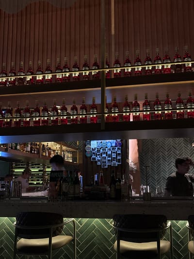The image shows a bar with rows of Campari bottles stacked on shelves. A customer sits at the bar, with a bartender visible behind the counter. A wall is decorated with a collage of photos. The bar top is made of a light-colored stone or marble, and the bar stools have a unique design. The background features a herringbone pattern, likely on the walls.