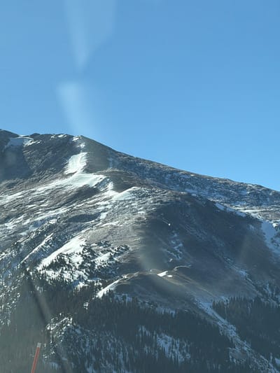 A steep, snow-dusted mountain face dominates the right side of the image against a clear blue sky. Jagged cliffs and rocky textures are highlighted by sunlight, creating dramatic shadows and depth. Patches of snow cling to the higher elevations, and some greenery appears on the lower parts of the mountain. A small red object is faintly visible on the bottom right side.
