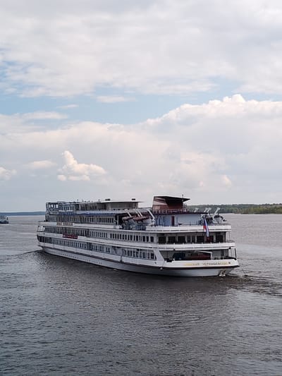 A medium shot depicts a cruise ship, named "Nikolay Chernyshevsky", sailing on a body of water. The ship is predominantly white with multiple decks. The name of the ship is written in gold lettering on the stern. A Russian flag is visible near the rear of the ship. The water is a dark gray color, showing signs of movement as the ship progresses. The sky is light blue with scattered white clouds. A strip of green land with trees can be seen in the background, suggesting a shoreline or riverbank. Another boat can be seen at a distance to the left of the main ship. No people are visible in the shot. Location is the city of Pruzhinino, Russia.