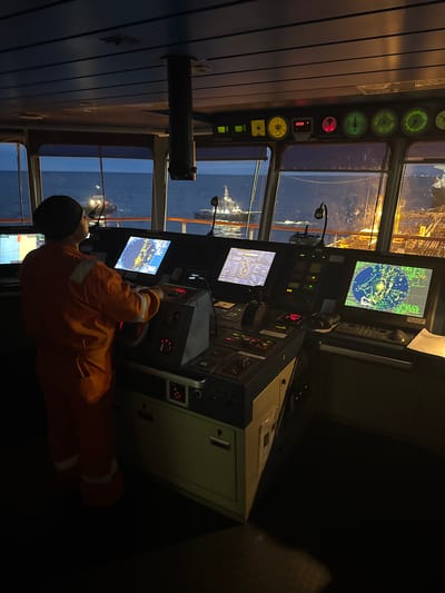 A person wearing an orange jumpsuit stands in the dimly lit bridge of a ship, facing a console filled with electronic equipment and screens. The person's back is to the camera, and they are looking at a large radar screen displaying maritime navigation data. Outside the windows, the sea is visible, with a tugboat in the distance and lights from another vessel on the right. The environment appears to be at sea during nighttime, indicated by the dark sky and the illuminated displays. The image does not contain any elements that suggest a location in Slobodka, Russia.