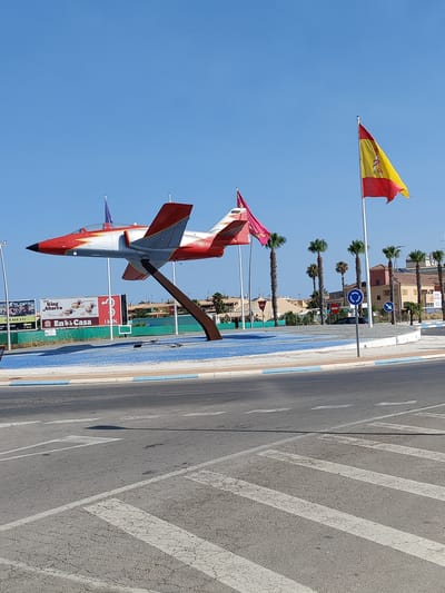 The image captures a monument of a red and white jet airplane, mounted on a brown support structure, situated in a roundabout in Los Alcázares, Spain. The jet is angled towards the left of the frame, with its nose pointing upwards. Two flags are visible; a Spanish flag flutters to the right of the jet, and another flag, possibly purple and blue, is placed on the jet itself. Surrounding the roundabout is a landscaped area with blue gravel and some vegetation. In the background, buildings and palm trees are visible under a clear blue sky. The foreground shows a street with road markings, including a pedestrian crossing. Advertisements are visible on a billboard behind the roundabout.