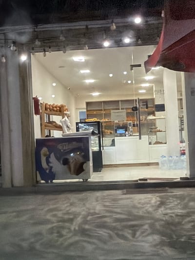 The image shows the exterior of a bakery, viewed through a glass storefront at night. The bakery appears to be well-lit from the interior, with various shelves displaying bread and pastries. A red awning is visible over the top right portion of the storefront. Inside, there is a counter area with what appears to be a cash register or display screen. Bottles of water are stacked on the floor to the right of the entrance. The foreground shows a reflective surface, possibly a wet pavement or the hood of a car. The location is specified as Tairov, Armenia.