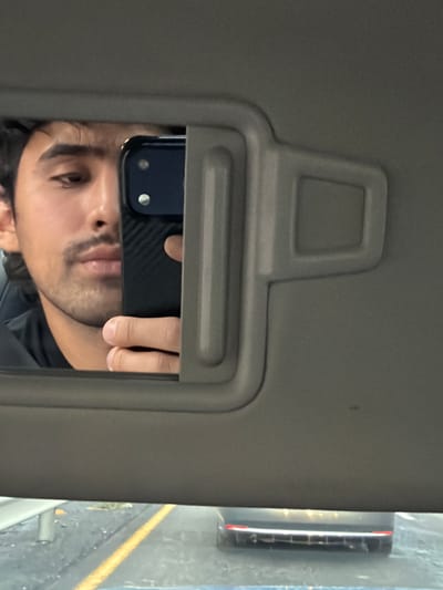 A selfie taken in the mirror of a car's sun visor. The person's face, hand and phone are reflected in the mirror. A car door is visible outside the car window. The location in San Bernardino Chalchihuapan, Mexico, cannot be determined from the image.
