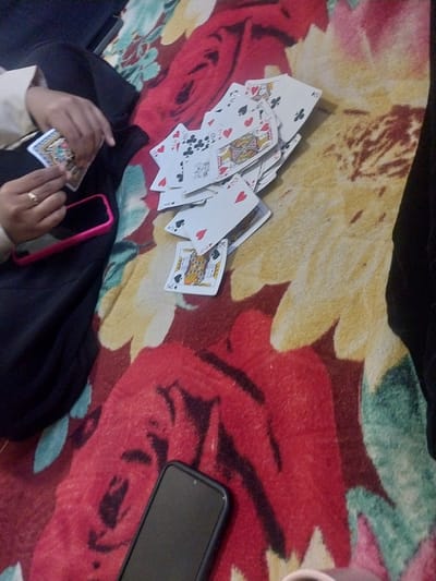 The image depicts a close-up view of a person's hands holding playing cards on a blanket with a floral pattern. The person is wearing a light-colored sleeve and dark pants, and a ring is visible on one finger. A pink smartphone is also present on the blanket. Several playing cards are fanned out, with visible suits and ranks including a King of Hearts, a 10 of Hearts, and a 4 of Clubs. The background is a heavily patterned blanket featuring large red and yellow floral designs. The lighting is dim. The scene is indoors. There is no indication of the city of Jeppee, Indonesia, or any specific geographical context beyond what is visible in the image.