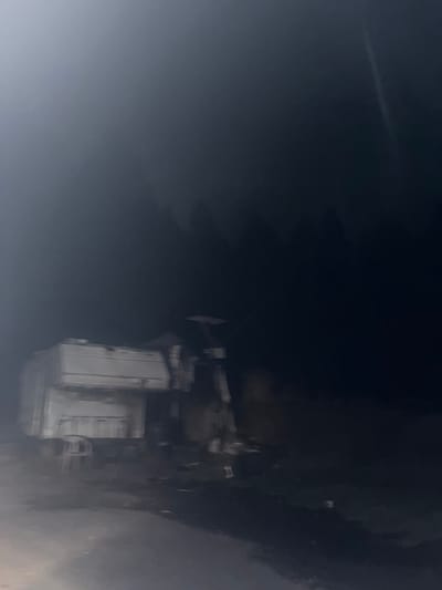 A partially obscured, unclear image appears to depict a building or structure at night. The building is white with doors visible. The right side of the image is dark, suggesting trees or shadows. The left side is gray and blurry, obscuring the view. A contrail is visible in the top-left corner.