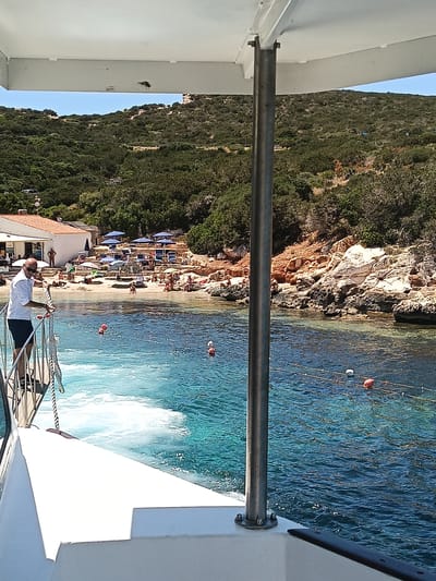 This is a photograph taken from a boat at sea looking towards a small beach with several people and a building. The boat is in the foreground, with a metal railing and a rope visible. The water is clear and blue, with buoys marking a swimming area. In the background, a sandy beach is lined with lounge chairs and umbrellas. A white building with an orange roof is situated behind the beach, with more lush green vegetation covering the hills beyond. The scene appears to be a sunny day in Alghero, Italy, likely a tourist destination.