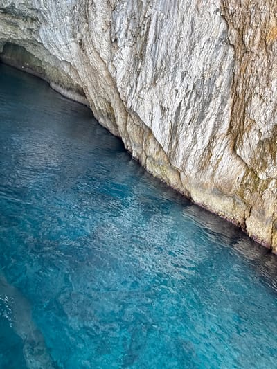 This image captures a close-up, diagonal view of a rocky cliff face meeting clear, turquoise water. The rocks are layered and textured, appearing weathered and ancient. The upper left section of the image is dominated by the rock formations, which range in color from pale, chalky white to golden brown, with visible striations and rough surfaces. A thin band of reddish-purple material, possibly algae or mineral deposits, runs along the edge where the rock meets the water.

The lower right portion of the image is filled with the ocean water. The water is a vibrant, translucent blue, with subtle ripples and variations in shade that suggest depth and movement. Sunlight refracts through the water, creating patterns of light and shadow. The overall impression is one of natural beauty and tranquility, typical of a coastal Mediterranean setting. There are no people or man-made objects visible, emphasizing the raw, natural landscape. The lighting suggests it is daytime, likely sunny, given the clarity of the water and the bright appearance of the rocks. The location context of Antipaxos, Greece, aligns with the visual cues of rugged coastline and clear blue waters.