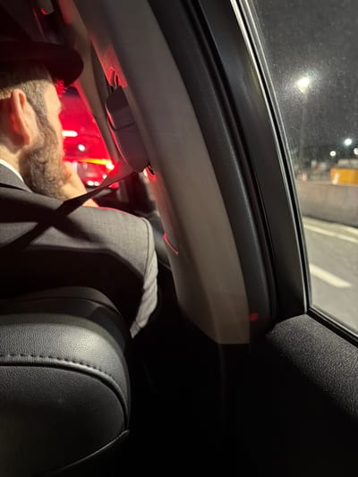 The photograph depicts the interior of a car at night, likely in traffic in Fort Lee, NJ. A person is visible in the driver's seat, seen from the back, wearing a dark suit jacket and a light-colored collared shirt. The person's face is turned upwards, with their ear and part of their beard visible. The driver is wearing a seatbelt. Red lights, possibly taillights, are visible outside the car's windows, illuminating the interior. The car has a sunroof, and the car's interior is dark, with the leather seats appearing black.