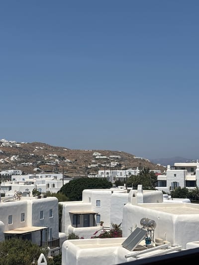 The image presents a daytime, panoramic view of Ornos, Greece, characterized by dominant white architecture and a clear sky. The upper portion of the frame is occupied by a cloudless, light blue sky. Below this, a large, arid, brown hill serves as the background, dotted with numerous white, cuboid structures and faint winding tracks. The mid-ground consists of a dense cluster of multi-story white buildings, typical of Cycladic architecture, featuring flat roofs, small rectangular windows, and occasional blue-painted shutters or frames. Interspersed among these buildings are areas of dark green vegetation, including at least one palm tree. The immediate foreground is composed of the white, flat rooftops of several structures. On a roof in the lower right quadrant, a solar water heater system is visible, comprising a horizontal cylindrical tank positioned above two angled solar collector panels. Other rooftops display vents and chimney-like features. No people, discernible actions, or interactions are present. The scene is brightly illuminated by direct sunlight, casting minimal shadows.